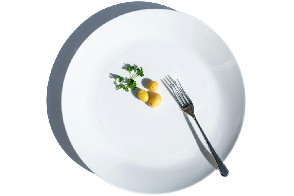 
طبق أبيض فارغ تقريبًا يحتوي على ثلاث حبات بطاطس صغيرة مع عود بقدونس وبجانبه شوكة – يرمز لتقليل الأكل أو الرجيم القاسي
